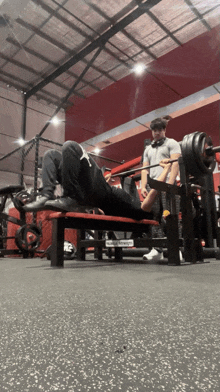 a man squatting on a bench in a gym with a man standing behind him with a barbell