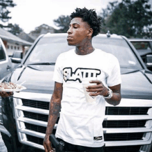 a young man in a white t-shirt is standing in front of a car holding a plate of food and a drink .