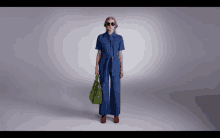 a woman wearing a denim jumpsuit and sunglasses holds a green bag