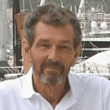 a man with a beard and a white shirt is standing in front of boats in a harbor .