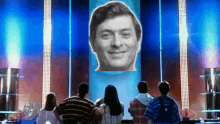 a group of people are sitting in front of a large screen with a man 's face on it