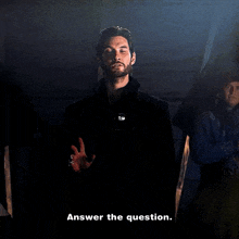 a man with a beard is standing in a dark room and says answer the question .