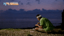 a man in a green shirt sits on the beach with a headband on his head and chopsticks in his hand