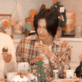 a man in a plaid shirt is sitting at a table with a christmas tree in the background and drinking from a cup .