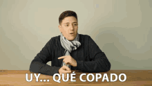 a man with a scarf around his neck is sitting at a table with the words uy que copado behind him