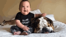 a baby is sitting on a bed next to a bulldog wearing a black shirt that says i 'm cute