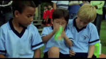 a group of young boys are sitting on the grass and one of them is drinking from a bottle .