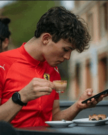 a man wearing a red shirt with a ferrari logo on it is drinking a cup of coffee