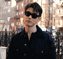 a man wearing sunglasses and a black jacket is standing in front of a fence with the letters nt behind him