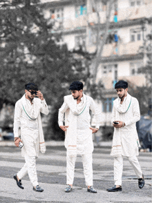 three men in white clothes are walking down a street and one is looking at his phone