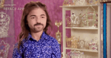 a man with a beard wearing a blue shirt is standing in front of a shelf filled with crowns .