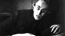 a black and white photo of a vampire laying on a bed looking at the camera .