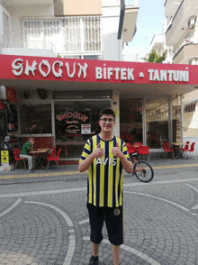 a boy wearing a yellow and blue striped shirt with avis on it stands in front of a restaurant