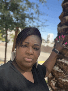 a woman in a black shirt and white earrings stands in front of a tree