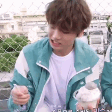 a young man in a blue jacket is eating ice cream with a spoon in front of a chain link fence .