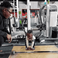 a man watches a little girl lift a barbell in a gym with a sign that says upped on the wall