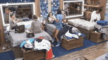 a group of women are sitting on beds in a messy room with clothes on the floor .