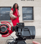 a woman in a red jacket is standing in front of a camera in front of a building that says box office