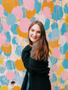 a woman in a black sweater stands in front of a wall with colorful spots