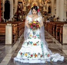 a bride in a white dress with embroidered flowers is standing in a church holding a bouquet of flowers