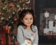 a little girl wearing a sweater with a snowman on it poses in front of a christmas tree and fireplace