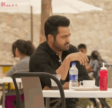 a man in a black shirt is sitting at a table with a bottle of water and a cup of coffee .