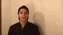 a young man wearing a black hat and a black jacket is standing in front of a white wall .