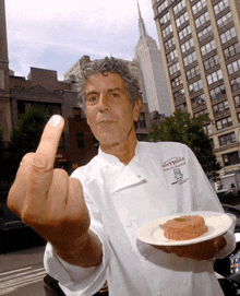 a man in a chef 's uniform holds a plate of food and a middle finger