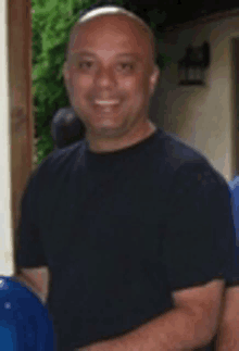 a bald man in a black shirt is smiling and standing in front of a building .