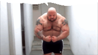 a bald man with tattoos on his arms and chest is standing in a hallway with his arms crossed .