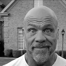 a bald man with a beard looks at the camera in front of a brick building