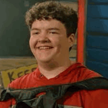 a young man in a red shirt is smiling while holding a black bag around his neck .