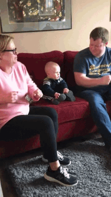 a baby sits on a red couch with a man wearing a blue shirt that says " best brand "