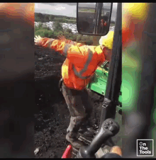 a man in an orange jacket and yellow hard hat stands in front of a green tractor with his arms outstretched ..