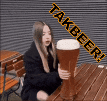 a woman is sitting at a table holding a large glass of beer with the words takbeer written above her