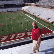 a man in a red shirt is standing on the roof of a stadium with a football field in the background