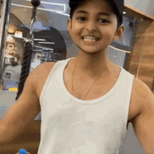 a young boy wearing a white tank top and a baseball cap is smiling while standing in a gym .