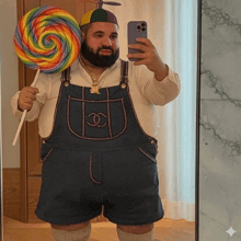 a man in overalls is holding a lollipop and taking a selfie with his phone