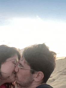 a man and a woman kissing on the beach with the man wearing glasses