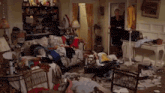 a woman is standing in a messy living room filled with lots of clothes and papers .