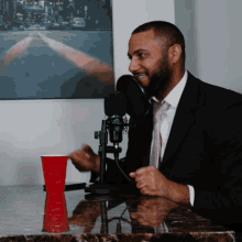 a man in a suit is sitting in front of a microphone with a red cup in front of him