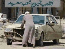 a man standing next to a broken down car with arabic writing on the wall behind him