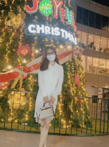 a woman in a white dress stands in front of a large christmas tree