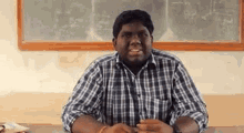 a man in a plaid shirt is sitting at a desk in front of a chalkboard in a classroom .