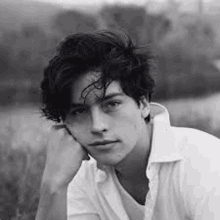 a young man in a white shirt is sitting in a field with his hand on his chin .