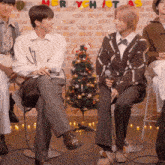 a group of people sitting in front of a christmas tree with merry christmas written on the brick wall behind them