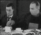 a black and white photo of two men sitting at a table with food . one of the men is pouring sugar into a cup .