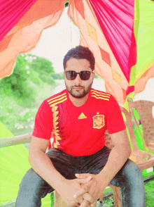 a man wearing sunglasses and a red adidas shirt sits in front of a tent