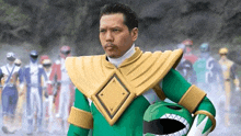 a man in a green and gold power ranger costume is holding a helmet in front of a group of power rangers .