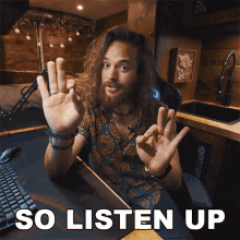 a man with long hair and a beard is sitting at a desk with the words so listen up behind him